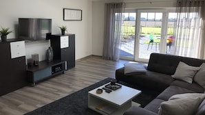 A modern living room with a sofa, a coffee table, a TV, and a sliding glass door leading to an outdoor patio. - Vacation apartments Königsgut Emmericher Eyland (apartment 2) (Kalkar)
