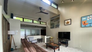 Ein geräumiges Wohnzimmer mit braunem Ledersofa, einem schwarzen Ledersessel, einem Holztisch, einem Fernseher und einem Deckenventilator.