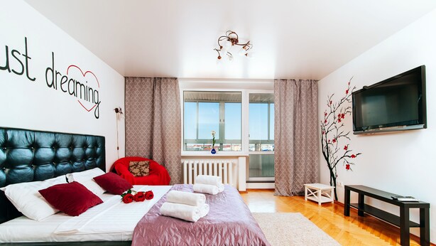 A modern bedroom with a large bed, a TV, and a balcony with a city view.