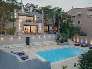 A modern two-story house with a swimming pool, outdoor seating, and a balcony with lights.