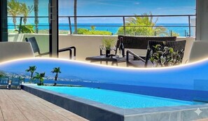 Una piscina a sfioro moderna con vista sull'oceano, sedie a sdraio e un tavolino con una pianta e una bottiglia.