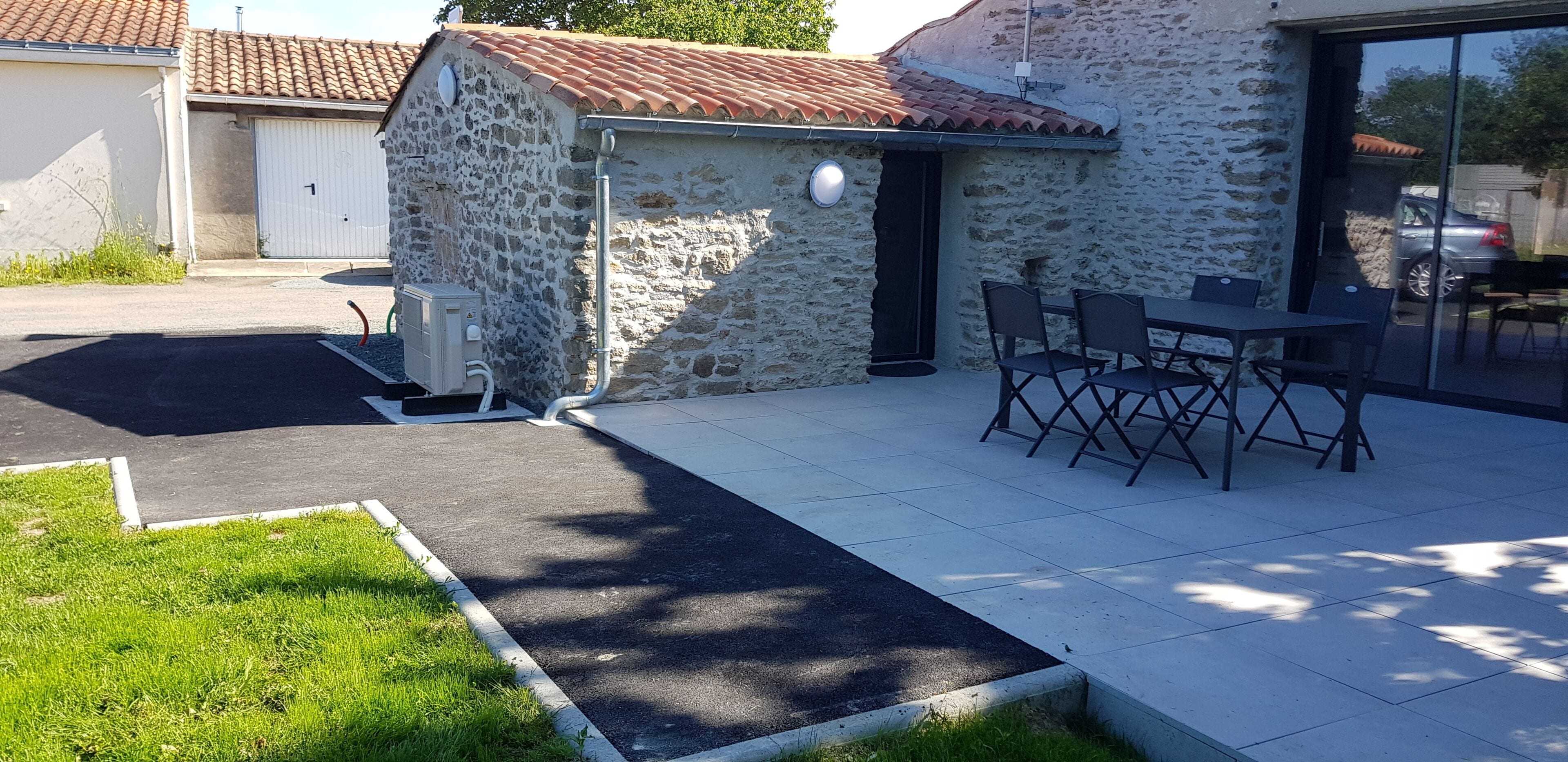 A stone building with a tiled roof, a small outdoor seating area with chairs, and a paved entrance.