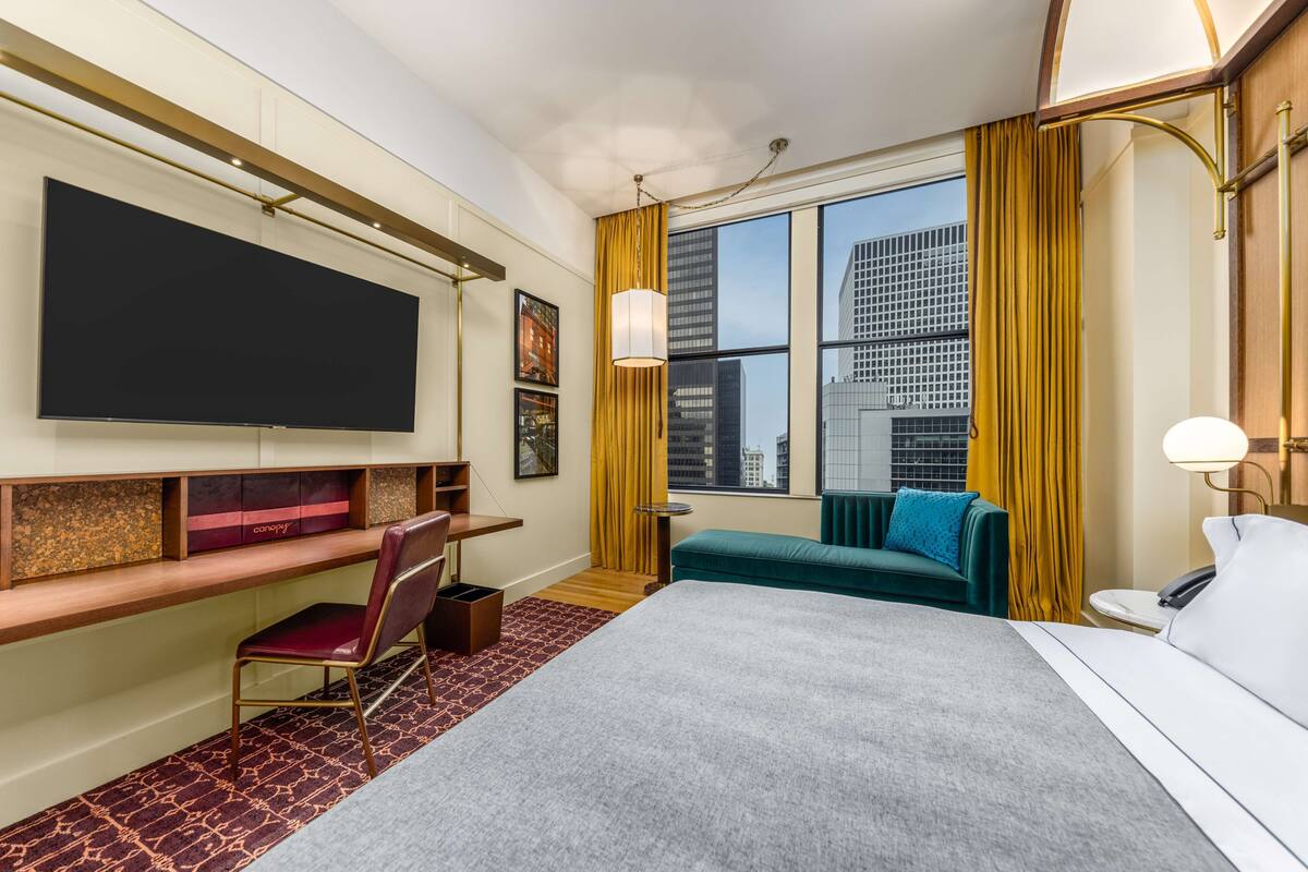 Habitación de hotel con cama, escritorio, silla, televisión y ventana con vista a la ciudad.