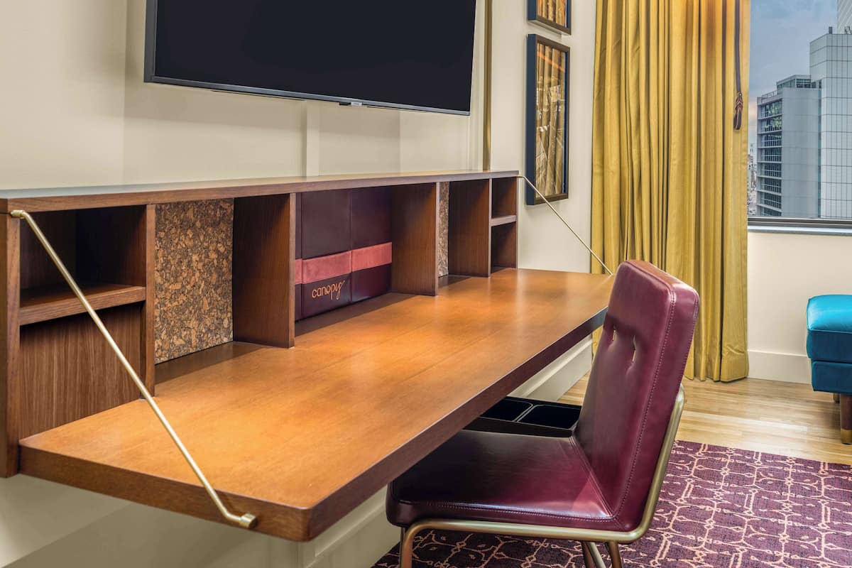 Habitación de hotel con escritorio integrado, televisor montado en la pared, ventana con cortinas y vista panorámica de la ciudad.