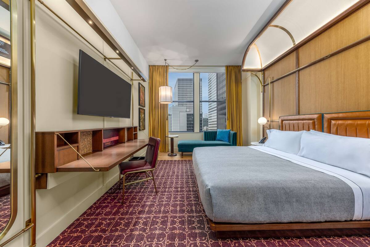 Habitación de hotel con una cama grande, un escritorio con silla, un televisor en la pared y vistas a la ciudad por la ventana.