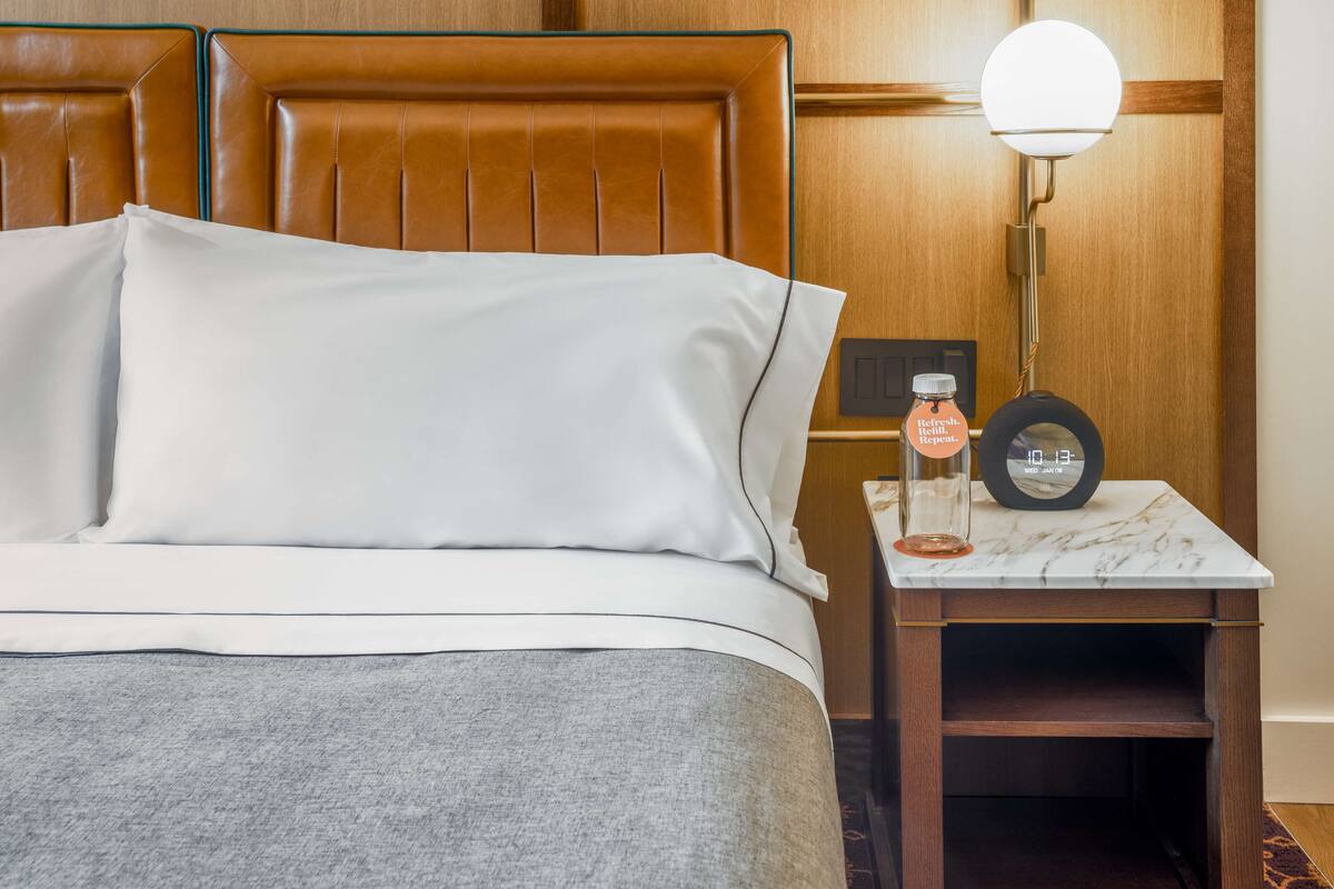 Una cama bien hecha con sábanas blancas, una mesita de noche con una botella y un reloj, y un cabecero de madera.