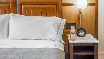 Una cama bien hecha con sábanas blancas, una mesita de noche con una botella y un reloj, y un cabecero de madera.