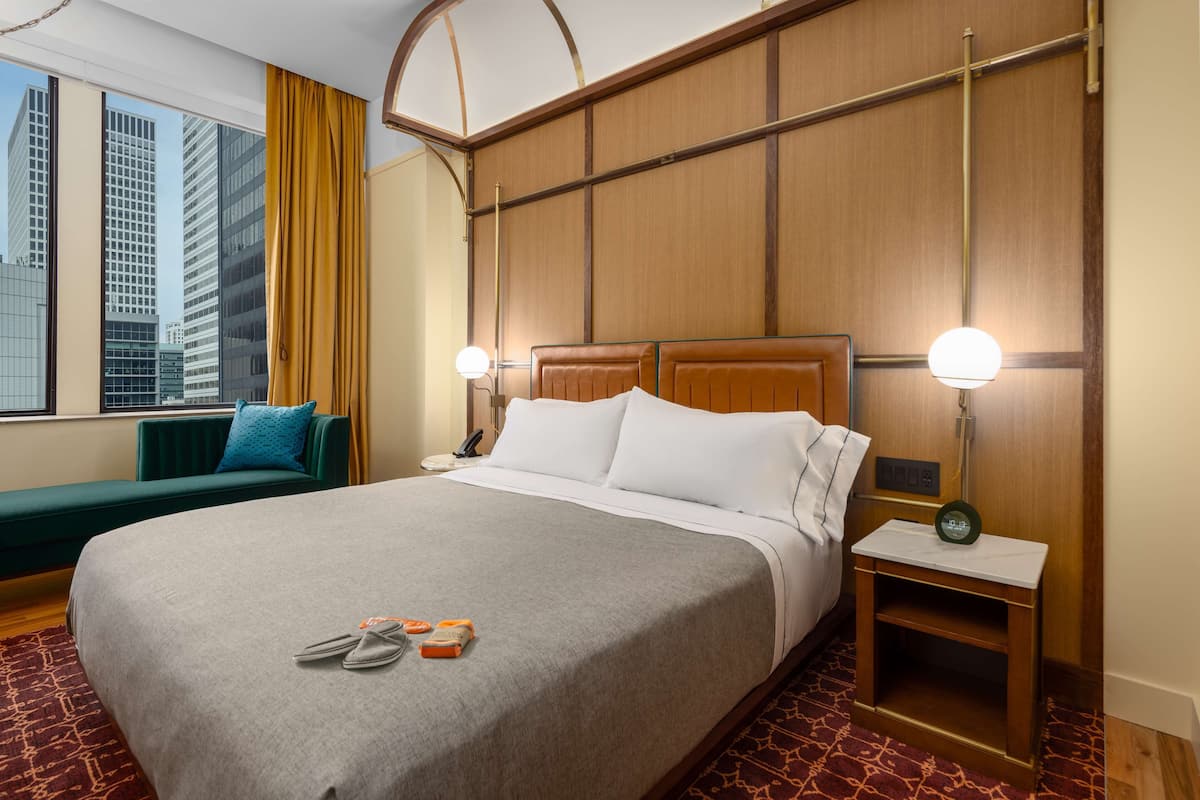 Habitación de hotel con una cama grande, una mesita de noche con lámpara, un sofá verde y vistas a edificios altos.