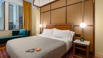 A hotel room with a large bed, a bedside table with a lamp, a green sofa, and a view of tall buildings.