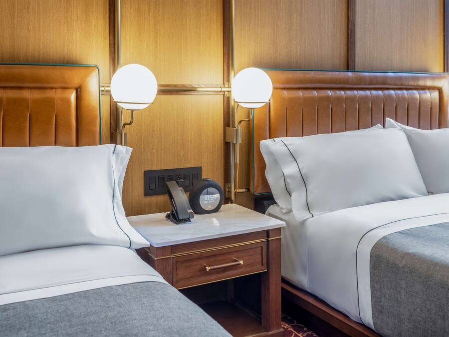 A hotel room with two beds, a bedside table with a clock, and wall-mounted lamps.