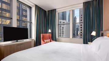 A hotel room with a large bed, a television on a wooden desk, a chair, and a view of city buildings through the windows.