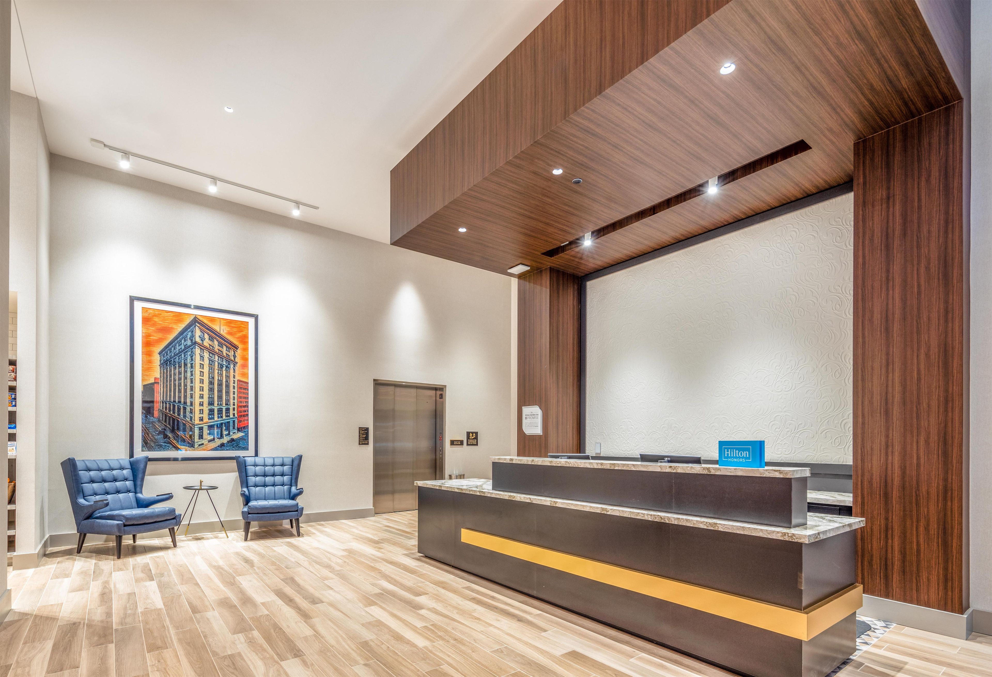 A hotel reception area with a reception desk, two blue chairs, a painting of a building, an elevator, and a wooden ceiling.