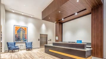 A hotel reception area with a reception desk, two blue chairs, a painting of a building, an elevator, and a wooden ceiling.