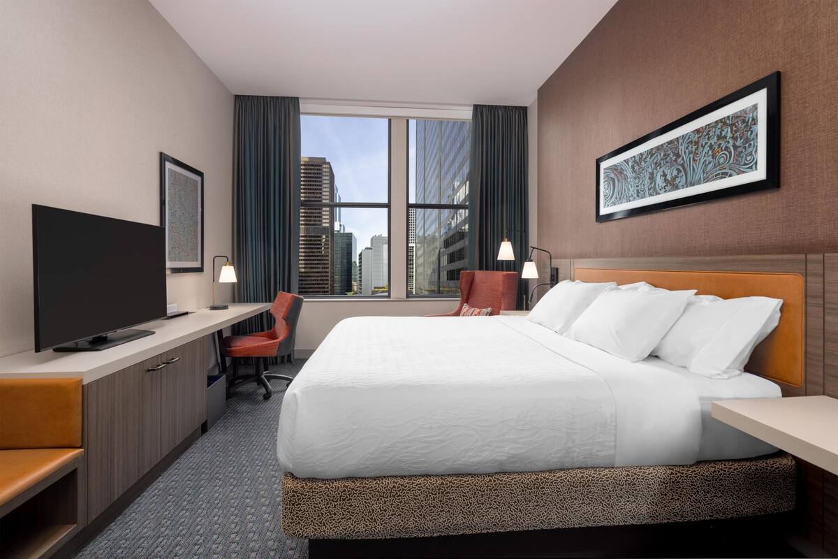 A hotel room with a bed, a desk, a television, a chair, and a window with a city view.