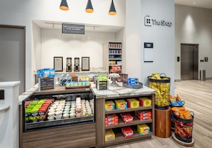 A snack display area with various packaged snacks, a coffee station, and a sign for 'The Shop' in a modern interior.