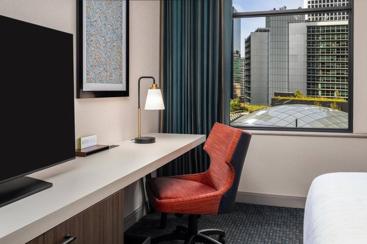 A hotel room with a bed, a desk with a lamp, a chair, a TV, and a window with a city view.