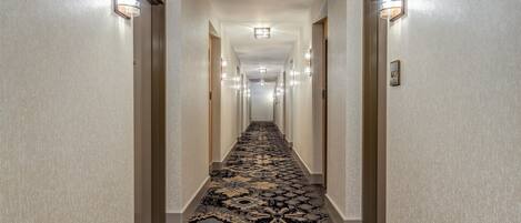 Pasillo de hotel con pisos alfombrados, luminarias fijadas a la pared y una puerta con placa de número.
