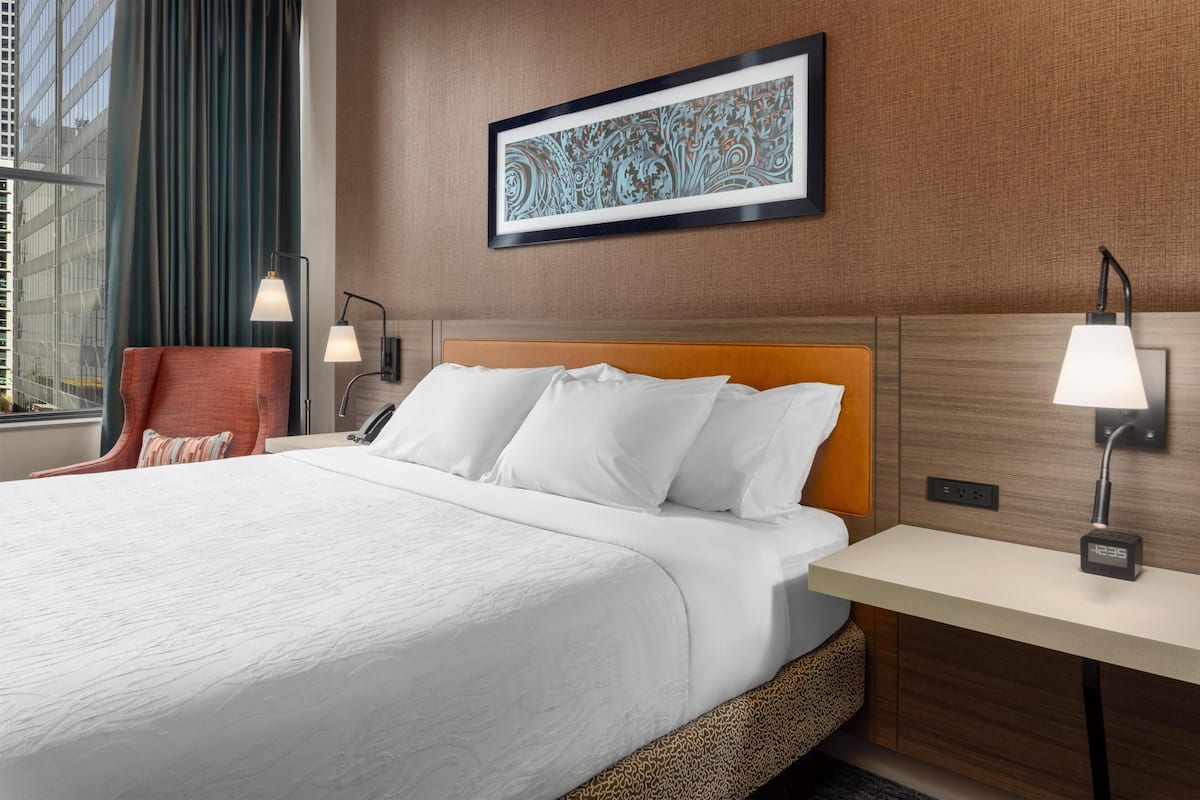A hotel room with a bed, a desk, a chair, a lamp, a framed artwork, and a window with curtains.