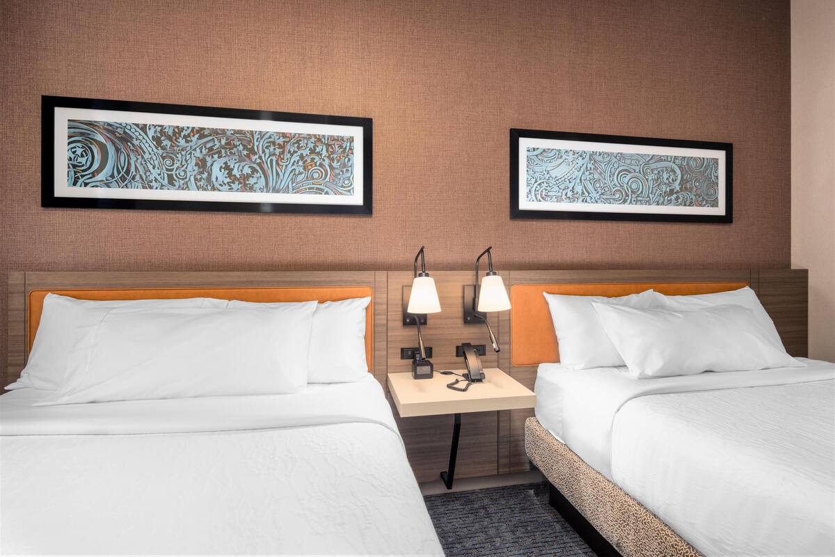 A hotel room with two beds, a nightstand with lamps, and two framed abstract wall art pieces.