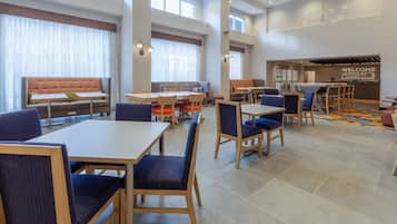 A hotel dining area with multiple seating options including tables and booths, a reception area with a 'Welcome' sign, and large windows providing natural light.