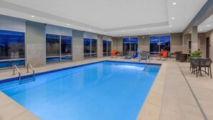 Une piscine intérieure à l’eau d’un bleu cristallin, entourée de chaises longues et de tables, de grandes fenêtres offrant une vue sur la ville et un aménagement intérieur moderne.