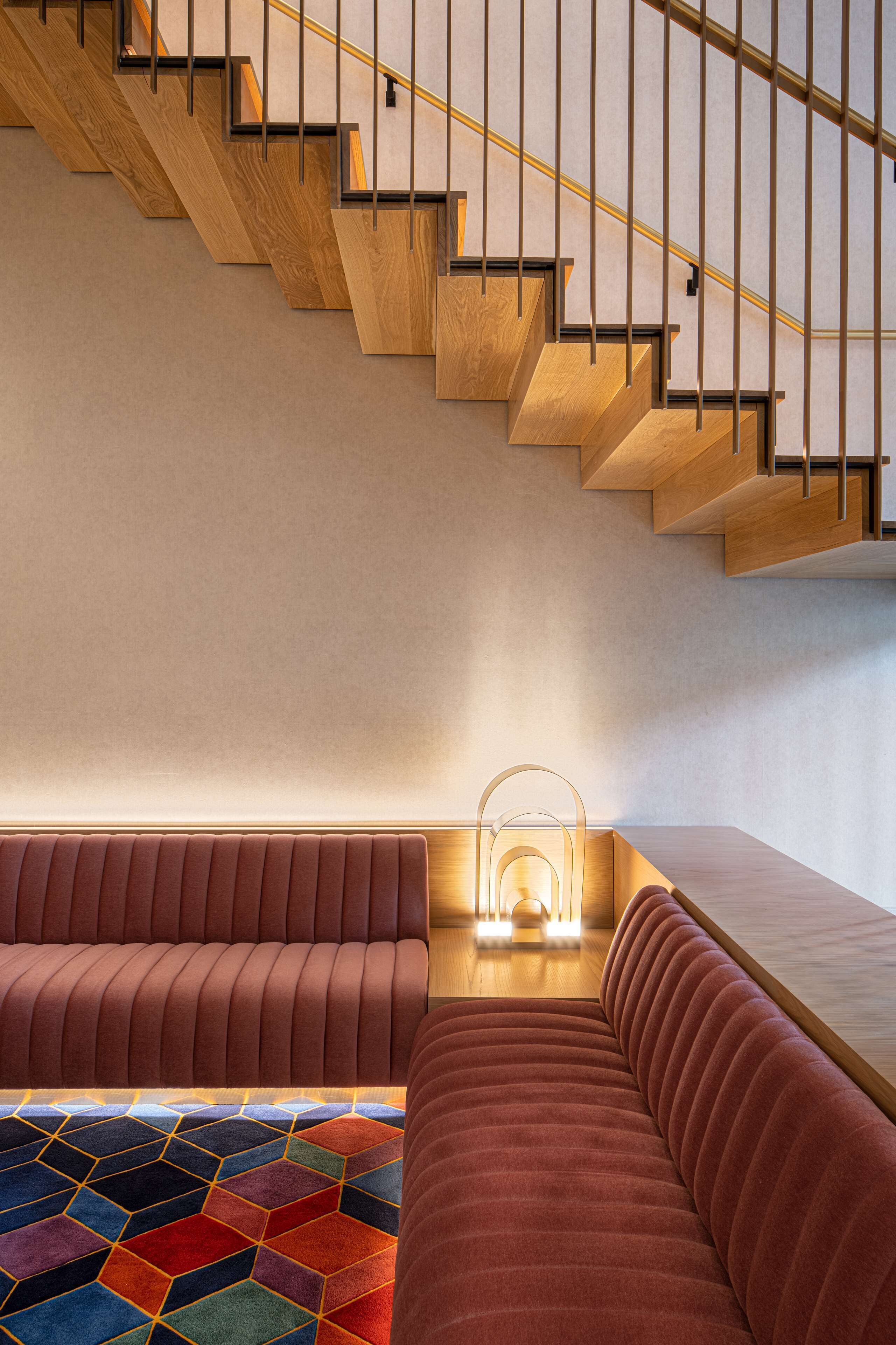 Un interior moderno con una escalera de madera, una zona de estar con alfombra estampada y una lámpara fijada a la pared.