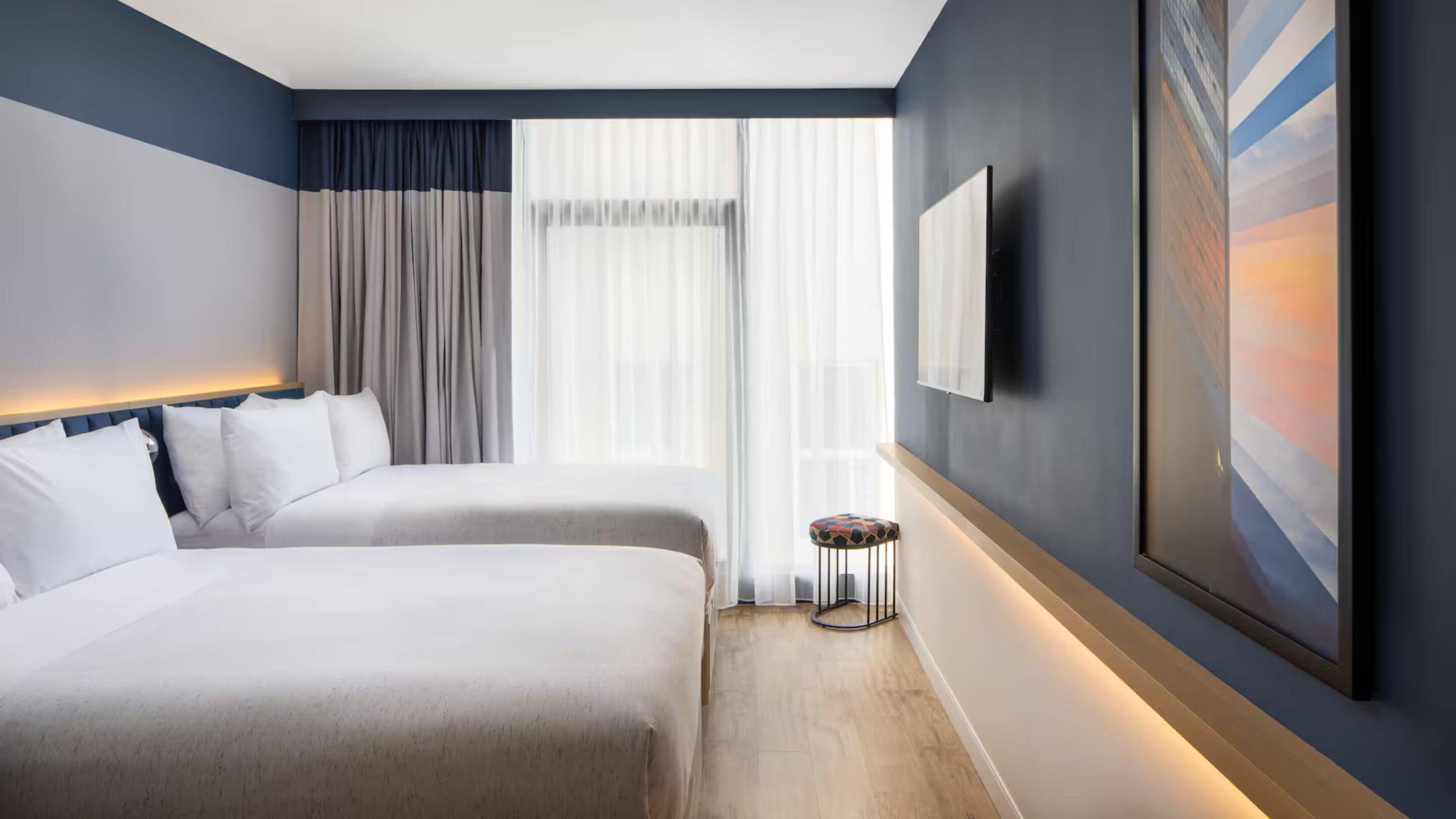 Camera d'albergo con due letti, una televisione, un quadro e uno sgabello.