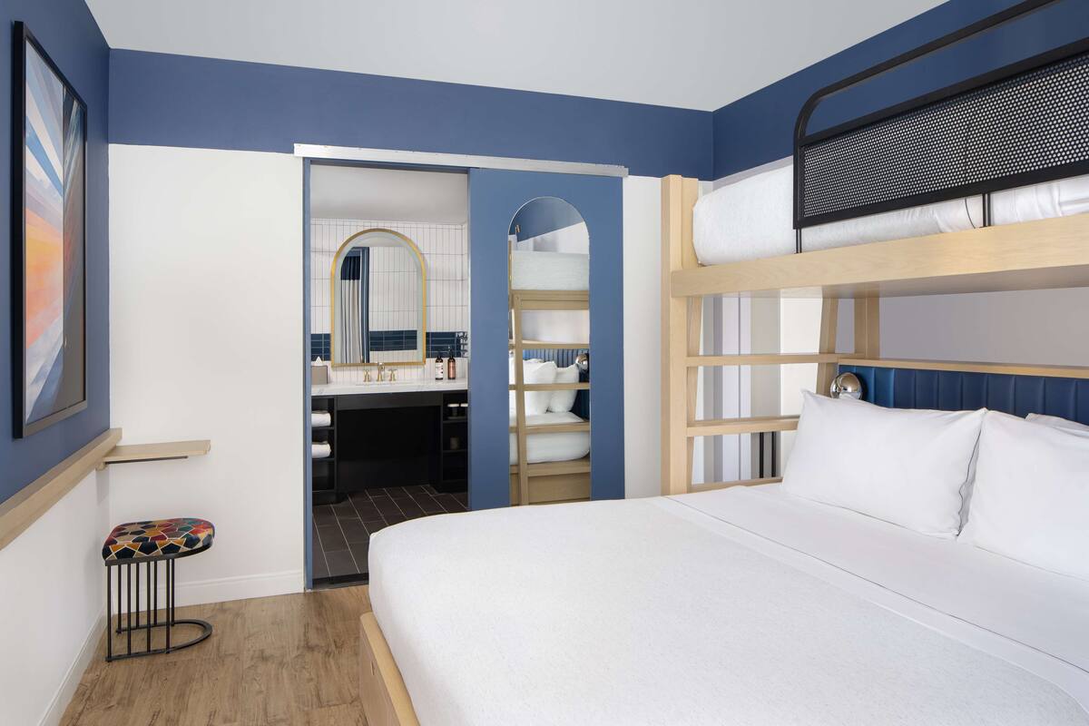 Ein Hotelzimmer mit Etagenbett, einem kleinen Hocker, einem Badezimmer mit Spiegel und einem gerahmten Bild an der Wand.