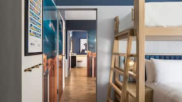 A hotel room with a bunk bed, a wooden ladder, a desk, and a chair. There is a hallway with framed pictures on the wall.