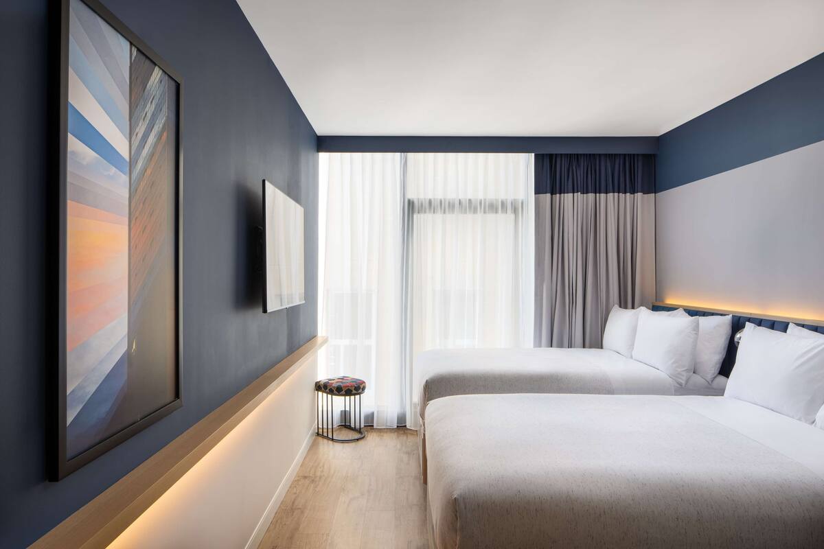Ein Hotelzimmer mit zwei Betten, einem großen abstrakten Gemälde an der Wand, einem kleinen Hocker und einem großen Fenster mit Vorhängen.