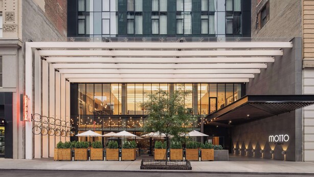 A modern building with a glass facade, outdoor seating area with umbrellas, and a visible restaurant interior. - Motto by Hilton New York City Chelsea (New York)