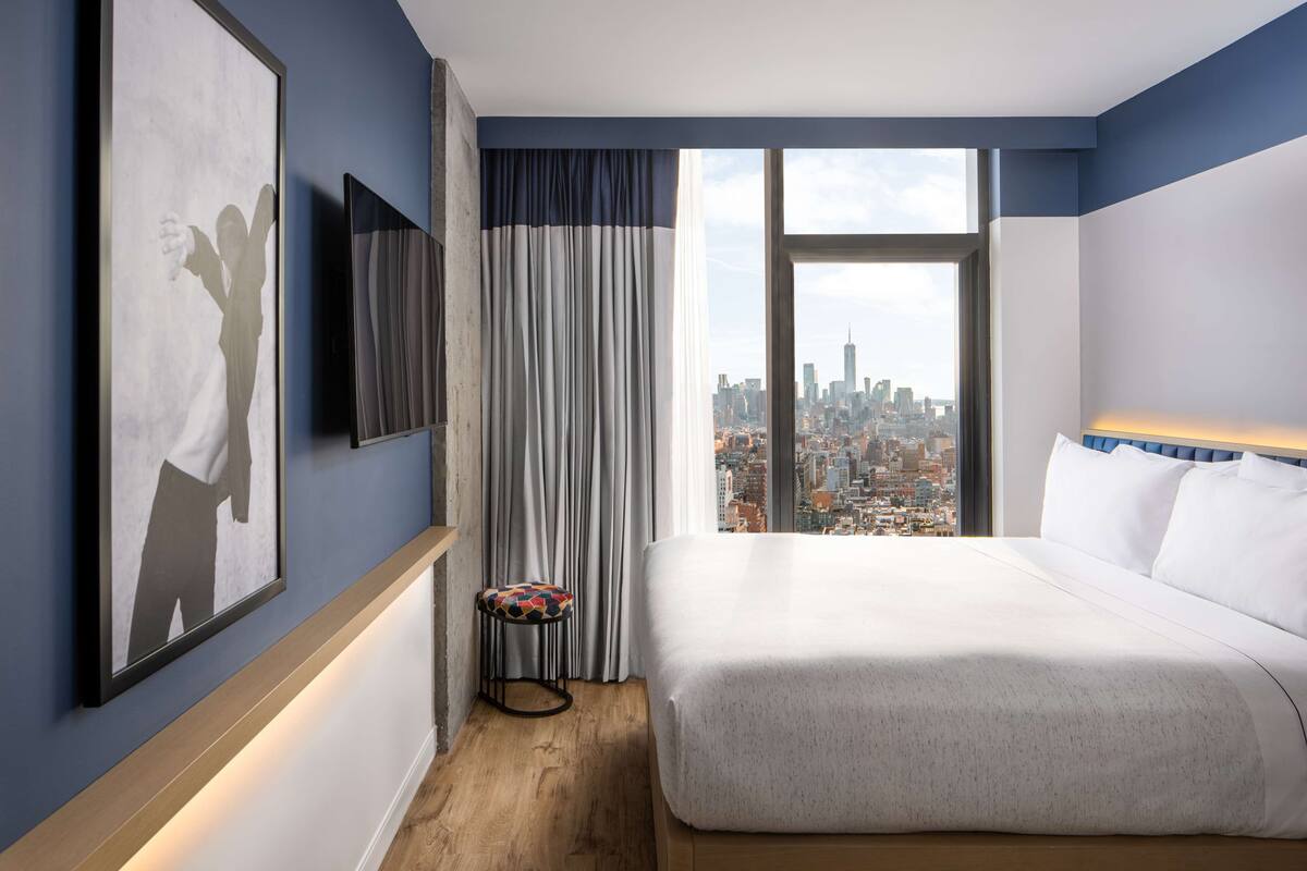Ein modernes Hotelzimmer mit einem großen Bett, Blick auf die Stadt, einem kleinen Hocker und einem gerahmten Bild an der Wand.