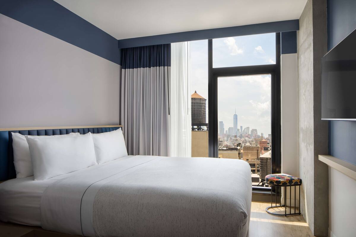 Ein Hotelzimmer mit einem großen Bett, Blick auf die Stadt durch ein großes Fenster und ein kleiner Hocker am Fenster.