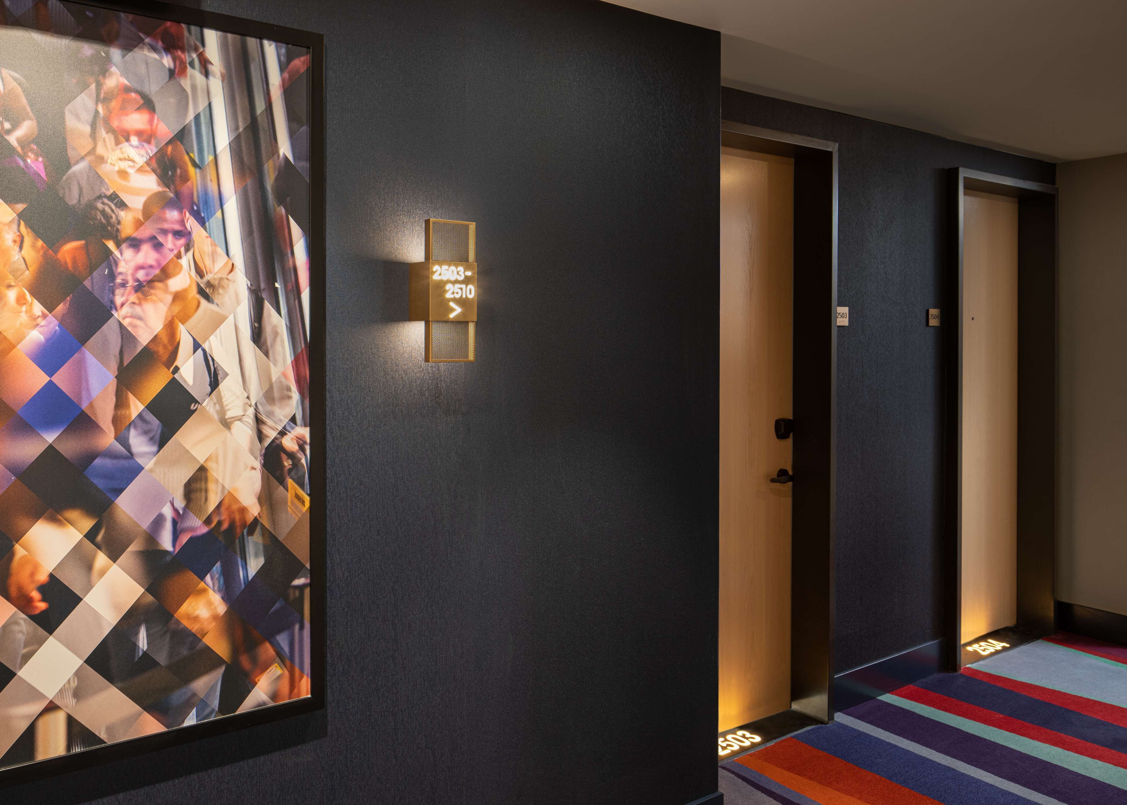 A hotel hallway with a large framed picture on the left, a sign indicating room numbers, and a colorful striped carpet.