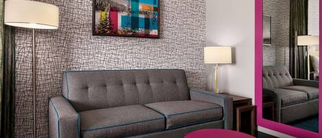 Una habitación de hotel moderna con un sofá gris, un otoman rosa, una lámpara de pie, una mesita auxiliar y un espejo grande.