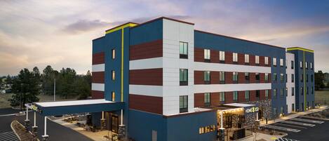 Un hotel de varios pisos con un diseño moderno, que presenta una combinación de colores azul, blanco y marrón. Cuenta con una entrada cubierta con un letrero, asientos al aire libre y un estacionamiento.