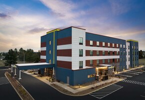 Un hotel de varios pisos con un diseño moderno, que presenta una combinación de colores azul, blanco y marrón. Cuenta con una entrada cubierta con un letrero, asientos al aire libre y un estacionamiento.