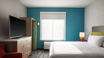 A hotel room with a bed, a TV on a wooden cabinet, a desk, a chair, and a window with blinds.