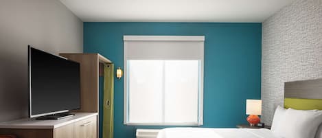 A hotel room with a bed, a TV on a wooden cabinet, a desk, a chair, and a window with blinds.