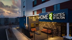 A hotel exterior with a sign reading "HOME 2 SUITES BY HILTON" and a patio area with seating. - Home2 Suites by Hilton Bend (Bend)
