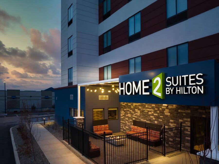 Exterior de un hotel con un cartel que dice "HOME 2 SUITES BY HILTON" y un patio con mobiliario.