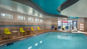 An indoor swimming pool with clear blue water, surrounded by yellow and white chairs, and a view of the outside through large windows. - Home2 Suites by Hilton Bend (Bend)