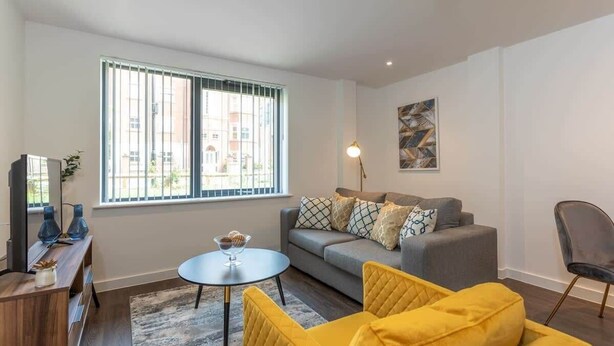 Een moderne woonkamer met een grijze bank, gele fauteuils, een ronde salontafel en een groot raam met zonwering.