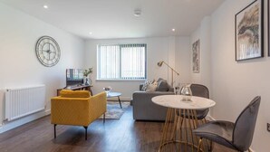 Een moderne woonkamer met een bank, een gele fauteuil, een ronde salontafel en een groot raam met zonwering.