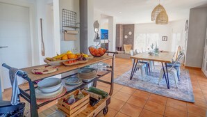 Uma cozinha com uma mesa de madeira, uma cesta de frutas e um carrinho com louças e utensílios.