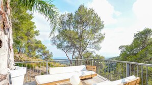 Um deck com bancos de madeira e almofadas brancas, rodeado por um guarda-corpo de metal, com vista para uma paisagem montanhosa.