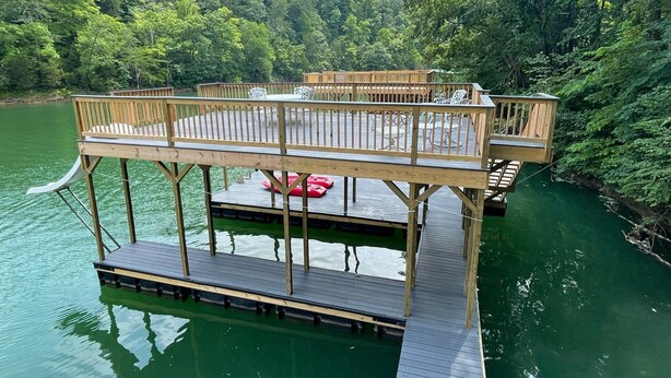 A wooden deck with seating and a small boat on a calm lake surrounded by dense forest.