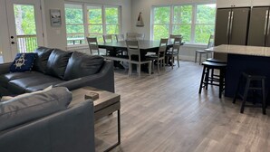 A spacious living room with a gray leather couch, a dining area with a large table and chairs, and a kitchen with a stainless steel refrigerator.