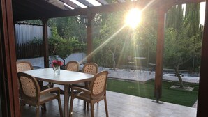 Une terrasse couverte avec une table et des chaises, une petite table avec une lampe, et une vue sur un jardin et des arbres.