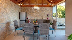 Een kamer met stenen muren en een houten plafond, een eettafel met rieten stoelen, een koelkast en een raam met uitzicht op bomen.